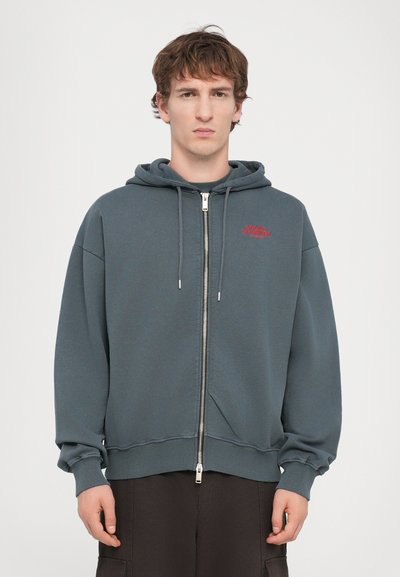 Grijze rits-hoodie met een ontspannen pasvorm, grote capuchon en rode "Snake Charmer" tekst. Beschikt over ribgebreide manchetten en een soepele textuur.