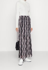 Kvinna klädd i en vit långärmad tröja, svart maxikjol med pastellfärgade paisleymönster, vita sneakers och hållande en svart handväska med kedjehandtag.