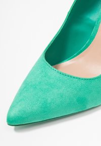 Chaussure en daim vert à bout pointu avec une doublure intérieure lisse et un léger éclat. Présente une forme élégante et des coutures nettes le long des bords.