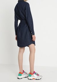 Jeanskjol med bälte i midjan, långärmad och med rundad nederkant. Kombineras med färgglada, klumpiga sneakers med grafiska detaljer.