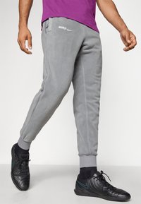 Sive trenirke s patentom na gležnjevih, s sivim flisom, ki vsebuje bel logotip z napisom "BARCA" in diskretnim Nike swooshom. Uporabljene z črnimi športnimi čevlji.