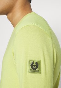 Pull vert clair avec un panneau d'épaule texturé et un patch logo Belstaff noir brodé sur la manche.