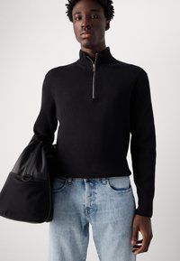 Crni rebrasti zip-up pulover s visokim ovratnikom, uparen s svijetloplavim trapericama, držan uz crnu platnenu torbu.