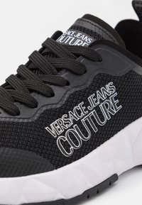 Svart textil sneakers med en strukturerad mesh-överdel, gummidetaljer, en djärv vit "VERSACE JEANS COUTURE" logotyp och en vit geometriskt mönstrad sula.