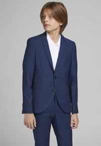 Costume ajusté bleu marine en tissu texturé, veste à un bouton, revers crantés et pantalon assorti. Chemise blanche à col en dessous.