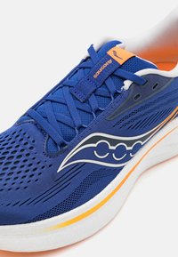 Chaussure de sport bleue avec une tige en maille respirante, accents orange, semelle extérieure blanche et un design de logo distinctif sur le côté.