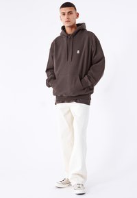 Mörkbrun hooded sweatshirt med en framficka, designad med en avslappnad passform. Kombinerad med lösa vita byxor och grå sneakers.