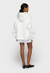 Sweat à capuche blanc surdimensionné avec une poche kangourou à l'avant et un cordon de serrage à la capuche, tissu doux, poignets côtelés et ourlet arrondi. Porté avec des shorts blancs.