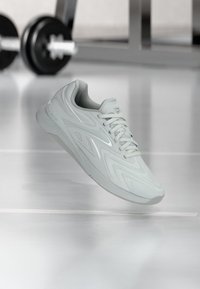 Αθλητικό παπούτσι ανοιχτού γκρι με άσπρα κορδόνια που αιωρείται πάνω από γυαλιστερό δάπεδο γυμναστηρίου, με θολό εξοπλισμό αλτήρων στο φόντο.