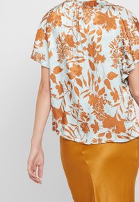 Kvinde iført en lyseblå bluse med orange blomsterprint og en sennepsgul satin nederdel, vist fra ryggen og siden.