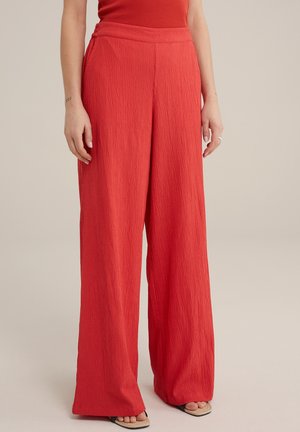 Femme portant un pantalon rouge taille haute à jambes larges et texturé, assorti à un top rouge et des sandales ouvertes, debout devant un fond neutre.