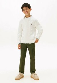 Suéter blanco de punto con cable, pantalones cargo verdes con bolsillos, zapatillas beige, postura de pie, expresión sonriente. Fondo sencillo.