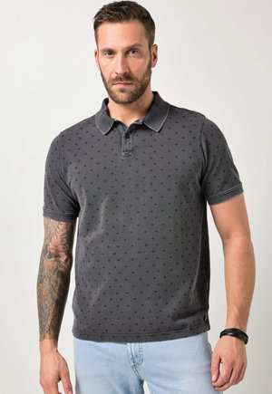 Mężczyzna z brodą i wytatuowaną ręką nosi ciemnoszarą koszulkę polo w kropki oraz jasnoniebieskie dżinsy, stoi na tle jednolitego jasnego tła.