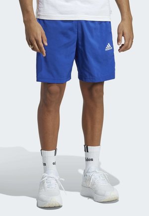 Mężczyzna w niebieskich szortach Adidas, białych skarpetkach Adidas i białych trampkach, stojący na jednolitym jasnym tle.