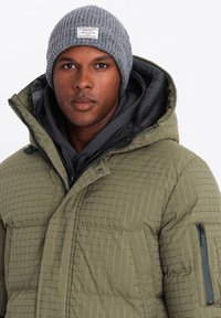 Chaqueta acolchada de color verde oliva con capucha, con un forro negro, bolsillo con cremallera y una sudadera gris en capas debajo. Gorro de punto gris en la parte superior.