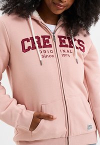 Felpa con zip di colore rosa chiaro, con un morbido rivestimento interno, grande testo ricamato in bordeaux, due tasche frontali e cappuccio con cordino.