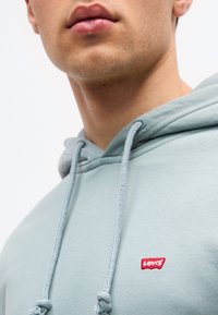 Μερική όψη ενός ατόμου που φοράει ένα ανοιχτό μπλε φο ύτερ Levi’s με κορδόνια και κόκκινο λογότυπο στο στήθος.