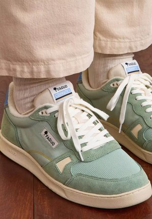 Green and beige Faguo Commute sneakers worn with beige socks and pants on a wooden floor.