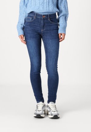 Osoba ubrana w dopasowane ciemnoniebieskie dżinsy, jasnoniebieski sweter z grubym splotem oraz białe masywne trampki, stojąca na jednolitym tle.