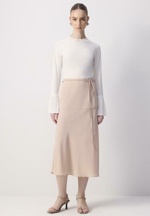 Long-sleeved white top with pleated cuffs paired with a knee-length beige skirt featuring a side tie and smooth texture. High-heeled sandals.