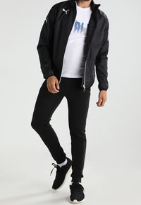 Chaqueta ligera negra con cierre de cremallera, logo blanco de Puma, combinada con pantalones negros ajustados y zapatos deportivos negros. Camiseta blanca de cuello redondo visible.