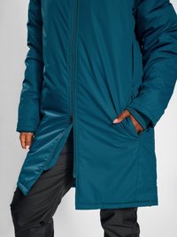 Long manteau matelassé de couleur sarcelle avec une fermeture éclair frontale partiellement ouverte en bas, porté sur un pantalon noir avec les mains dans les poches du manteau.