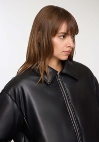 Młoda kobieta z jasnobrązowymi włosami, ubrana w czarną skórzaną kurtkę na zamek, patrzy na bok na jednolitym jasnym tle.