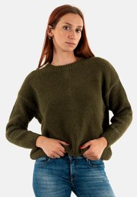 Oliven grøn, oversized strikket sweater med ribbet tekstur, rund halsudskæring og dropped skuldre. Parret med blå jeans, hænderne på hoften.