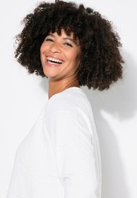 Sudadera blanca con un patrón texturizado, cuello redondo y corte relajado. El modelo tiene el cabello rizado y está sonriendo frente a un fondo blanco.