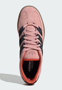 Rozā zamša sporta apavi ar melniem svītrainiem akcentiem, teksturētām auklām un gumijas zoli. Iekšpusē ir oranžas detaļas un logotips.