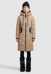 Beige puffer coat with chevron stitching, zip closure, and adjustable hood; features black pockets and accents; worn with black boots.