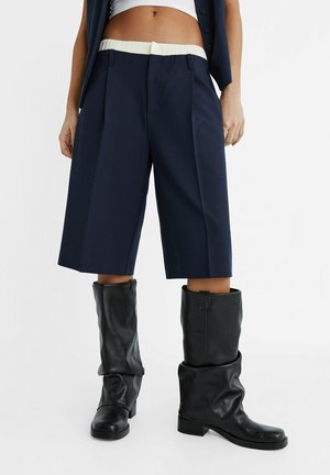 Personne portant un short bleu marine jusqu'aux genoux avec une ceinture élastique blanche, des bottes en cuir noires amples, un top blanc court et un gilet bleu marine.