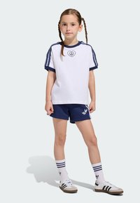 Biele tričko s námorníckymi detailmi a srdcovým logom, kombinované s námorníckymi šortkami. Biele ponožky s modrými pruhmi a čierno-biele tenisky dotvárajú vzhľad.