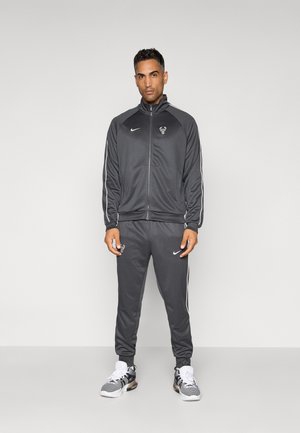 Grå atletisk træningsdragt med hvide sidelister, der har et logo på brystet og på buksebenet. Stoffet virker glat og let.