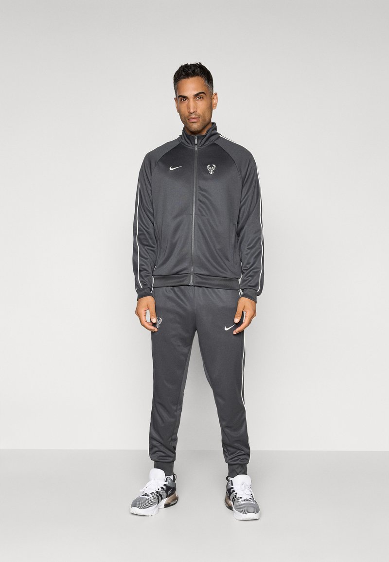 Gray athletic tracksuit with white side stripes, featuring a logo on the chest and pant leg. Fabric appears smooth and lightweight.
