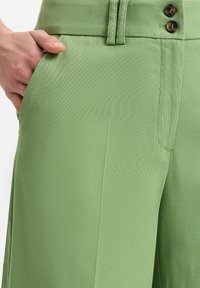 Grüne Hose aus einem glatten, strukturierten Stoff, mit zwei braunen Knöpfen und genähten Details am Bund und den Seitentaschen.