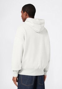 Uomo visto di spalle che indossa un felpa con cappuccio grigio chiaro e jeans blu scuro su uno sfondo bianco uniforme.