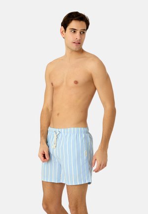 Junger Mann steht oberkörperfrei und trägt hellblaue Badeshorts mit weißen vertikalen Streifen und einem weißen Kordelzug.