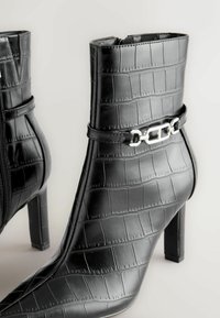 Bottines noires en simili-cuir crocodile texturé, avec un accent en chaîne argentée, un talon haut et une fermeture éclair sur le côté.