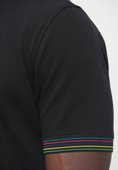 Detailný záber na hornú časť ramena osoby, ktorá má na sebe čierne tričko s krátkym rukávom, zdobené viacfarebným pruhovaným lemom na okraji rukávu.
