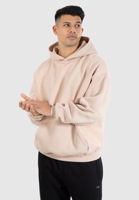 Haut à capuche beige clair avec une coupe décontractée, grande poche frontale et capuche réglable par cordon. Possède des poignets et un ourlet côtelés. Pantalons de survêtement noirs.