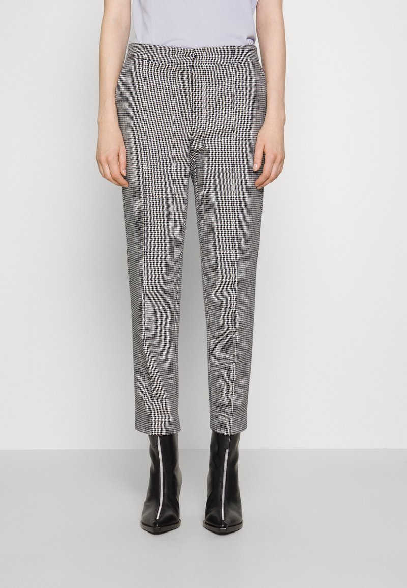 Personne portant un pantalon slim à carreaux et des bottines noires à bout pointu avec une fermeture éclair sur le devant, debout devant un fond clair uni.