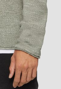 Hellgrüner Strickpulloverstoff mit einem strukturierten Muster. Verfügt über einen umschlagenen Ärmel und eine schlichte weiße Innenschicht, die am Handgelenk sichtbar ist.
