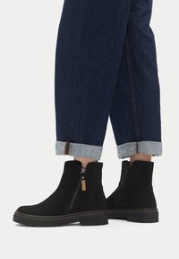 Botines de gamuza negra con cremallera lateral, con detalle de costuras en contraste y suela de goma resistente, combinados con vaqueros con los bajos remangados.