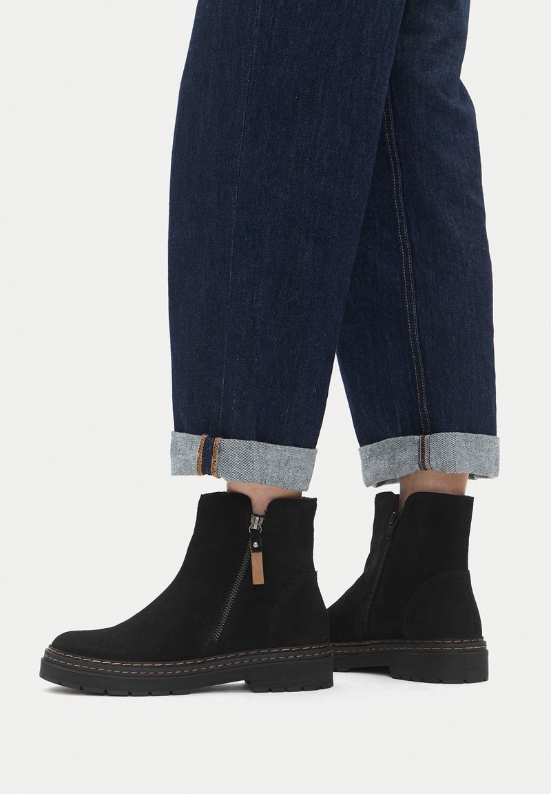 Botines de gamuza negra con cremallera lateral, con detalle de costuras en contraste y suela de goma resistente, combinados con vaqueros con los bajos remangados.