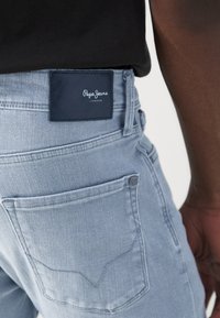 Svijetloplave traperice s teksturiranim završetkom, koje sadrže tamnoplav kožnu oznaku na pojasu s natpisom "Pepe Jeans London" i stražnjim džepovima.