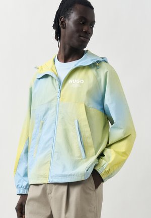 Νέος άνδρας που φοράει μια παστελ γαλάζια και κίτρινη ζακέτα με φερμουάρ και κουκούλα, με το λογότυπο "HUGO", μπεζ παντελόνι, στέκεται με τα χέρια στις τσέπες και κοιτάζει πλάγια.