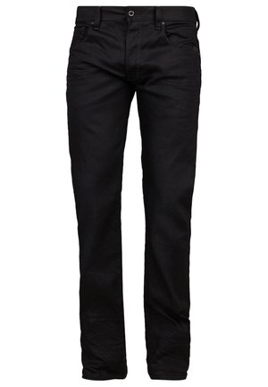 Mand iført sort jakke, sorte bukser, sorte sko, der holder en sort rygsæk ved sin side mod en ensfarvet baggrund.