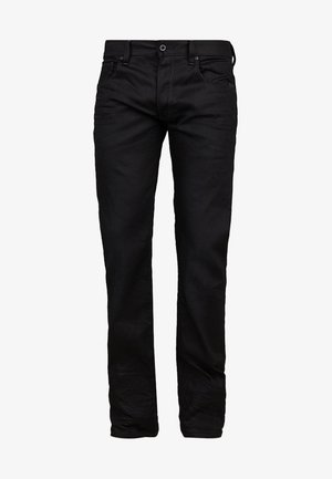 Man die een zwart jack, zwarte broek, zwarte schoenen draagt en een zwarte rugzak naast zich houdt tegen een effen achtergrond.