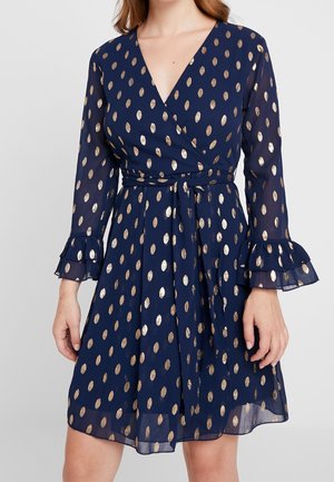 Marineblaues Wickelkleid mit langen, durchsichtigen Ärmeln, goldenen Polka Dots, Rüschenmanschetten und einem gebundenen Taillengürtel an einer stehenden Frau.